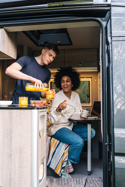 Young man serving natural juice to cheerful black girlfriend while enjoying breakfast together inside camper trailer during summer holidays — Stock Photo