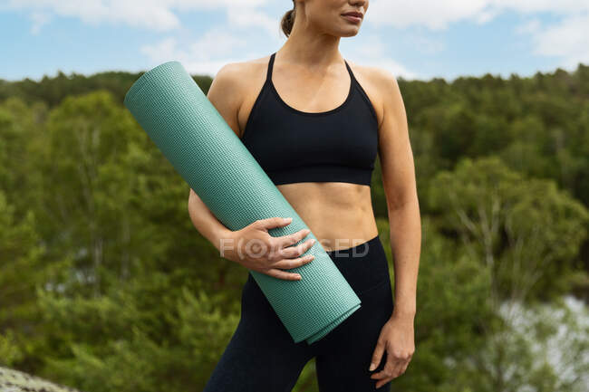 Mujer joven anónima sin emociones en ropa deportiva negra llevando estera enrollada y mirando hacia otro lado antes de la sesión de yoga en el exuberante campo - foto de stock
