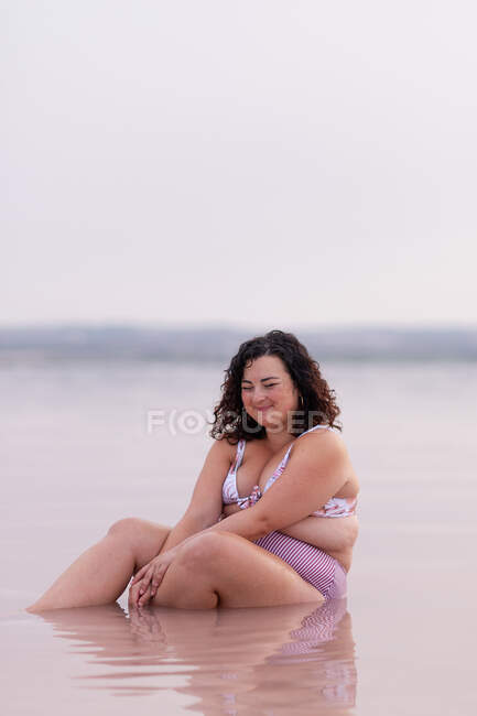 Восхитительная пышная женщина в бикини, сидящая в воде розового пруда летом и смотрящая вниз — стоковое фото