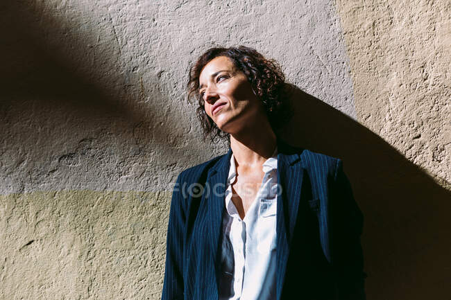 Задумчивая женщина в элегантной одежде стоит солнечный свет и отбрасывает тень на потрепанную стену и смотрит в сторону — стоковое фото