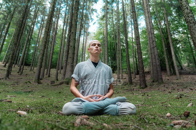 Uomo calvo in abiti tradizionali seduto sull'erba in posa Lotus e meditare durante la formazione di kung fu nella foresta — Foto stock