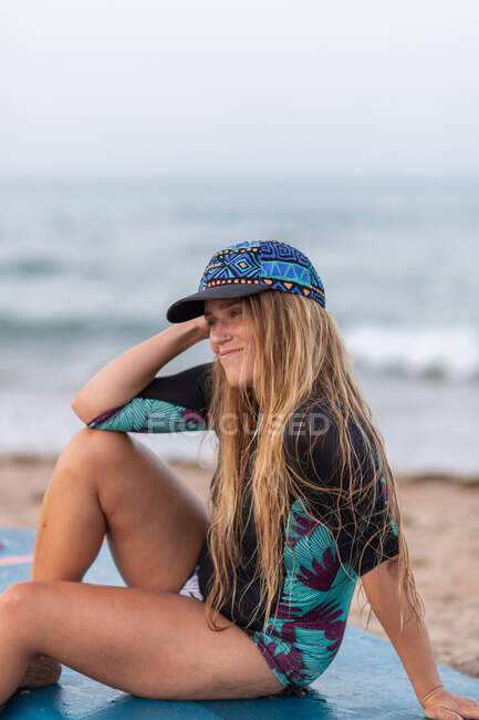 Side view of positive female surfer in swimwear and hat sitting on paddleboard on sandy shore against sea and looking away — Stock Photo