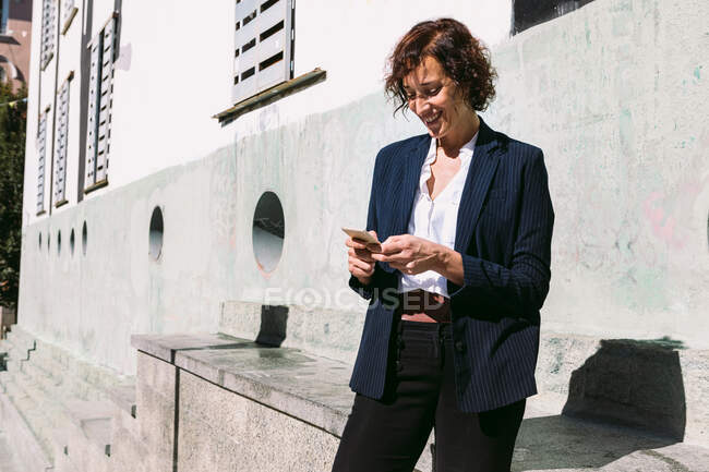 Positive adult female executive worker wearing classy outfit standing browsing on cellphone in sunny day — Stock Photo