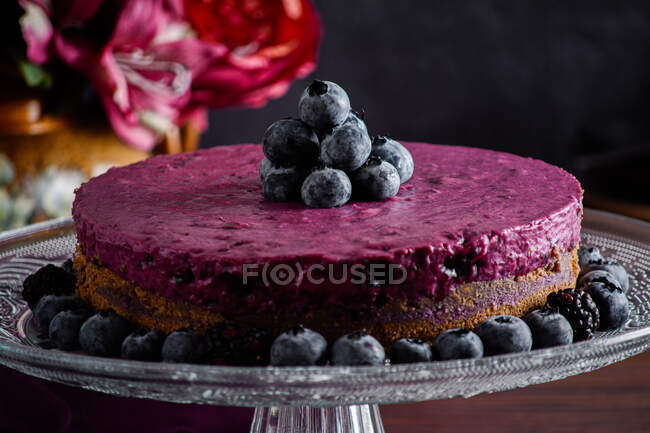 Вкусный черничный торт из мусса с фиолетовым кремом, украшенный свежими ягодами, подаваемый на стеклянном столике на темном столе с цветами — стоковое фото