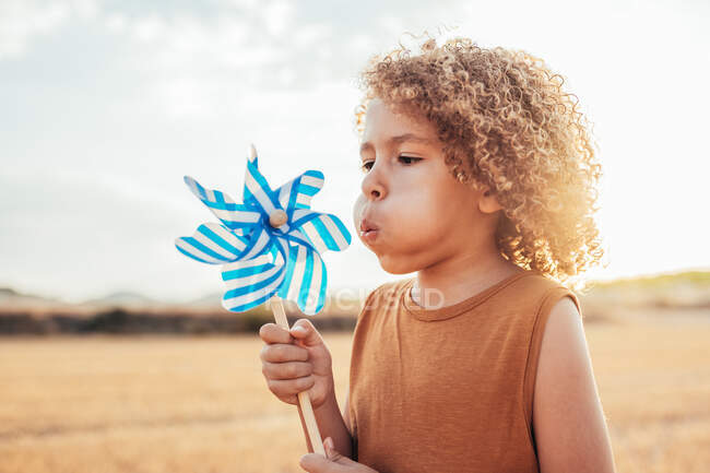 Ethnic kid with curly hair blowing on pinwheel while playing in field in countryside in summer — Stock Photo