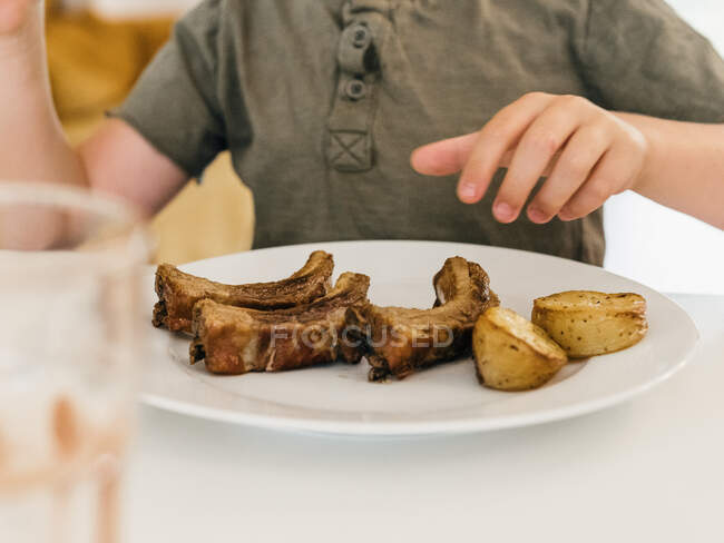 Crop of anonymous kid eating appetizing pork ribs during lunch at home — Stock Photo