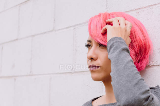 Pensive young female with dyed hair in casual clothes looking away while standing near white wall with his hand on his head — Stock Photo