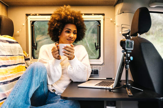 Cheerful young African American female traveler drinking coffee and enjoying video chat via laptop while resting inside camper van during summer adventure — Stock Photo