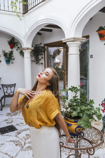 Anmutige Frau in stylischer Sommerkleidung steht neben Tisch mit Blumen im Innenhof des Hauses — Stockfoto