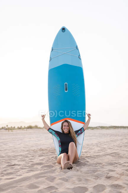 Fröhliche Surferin sitzt im Sommer mit blauem SUP-Board am Sandstrand und blickt in die Kamera — Stockfoto