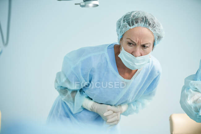 Концентрированная зрелая женщина-врач в стерильной форме наклоняется вперед, сжимая руки, глядя в операционную. — стоковое фото