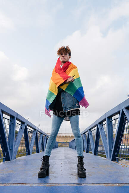 Desde abajo de un hombre homosexual serio de pie con la bandera del arco iris en el puente y mirando a la cámara - foto de stock