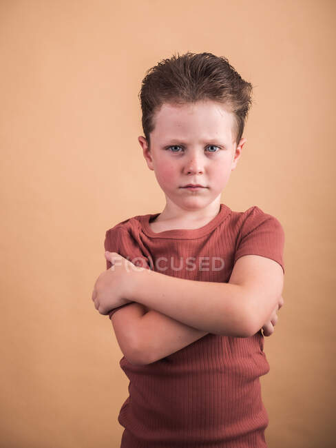 Неприємна дитина зі складеними руками і коричневим волоссям дивиться на камеру на бежевому фоні — стокове фото