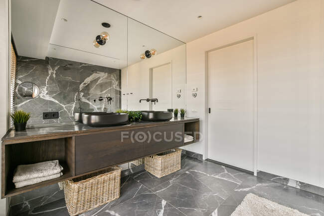 Elegante diseño interior de lujoso cuarto de baño espacioso con azulejos de mármol amueblado con lavabo doble y gabinete de madera marrón oscuro bajo un gran espejo en apartamento moderno - foto de stock