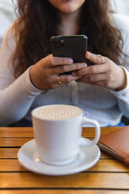 Обрізаний невпізнаваний етнічний жіночий обмін повідомленнями на мобільному телефоні, сидячи за столом з чашкою кави і блокнота в кафетерії — стокове фото