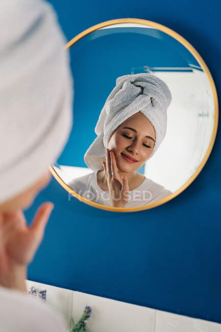 Visão traseira da fêmea em turbante de toalha refletindo no espelho no banheiro e aplicando creme facial durante a rotina de cuidados com a pele de manhã — Fotografia de Stock