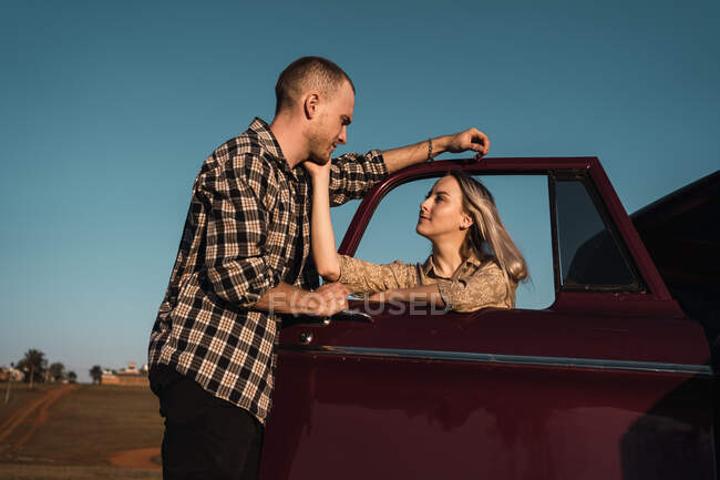 Seitenansicht einer liebenden Frau, die das Gesicht eines Mannes durch das geöffnete Fenster eines in der Natur geparkten Retro-Autos berührt — Stockfoto