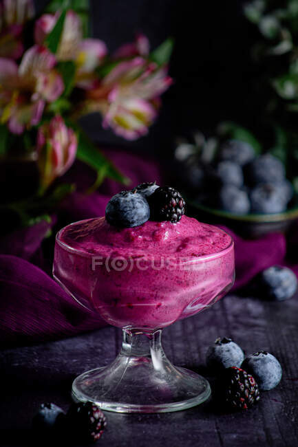 Glas mit frischem Blaubeermousse garniert mit frischen Beeren, serviert auf dunklem Tisch mit verschwommenen Blüten — Stockfoto