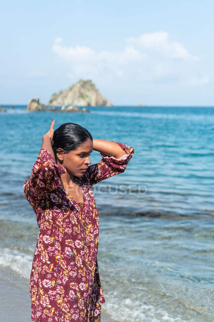 Mulher viajante étnica em vestido de verão com ornamento floral olhando para baixo com as mãos atrás da cabeça contra o oceano e a rocha — Fotografia de Stock