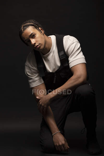 Side view of confident young ethnic hipster male model with braided hair wearing white shirt with black clothes looking at camera while crouched in dark studio — Stock Photo
