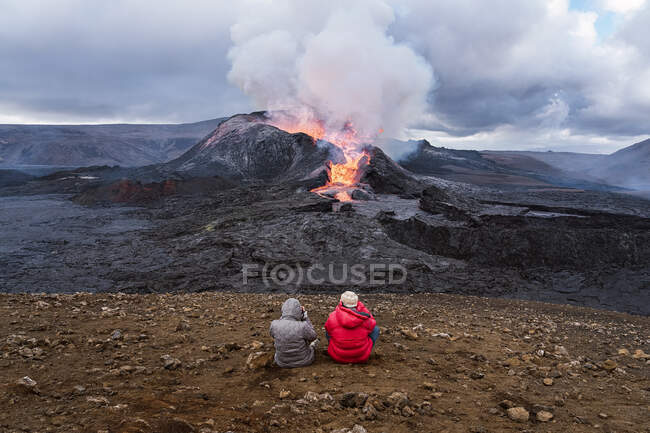 Vue arrière de voyageurs méconnaissables admirant Fagradalsfjall avec le feu et la lave tout en prenant des photos et assis sur le mont en Islande — Photo de stock