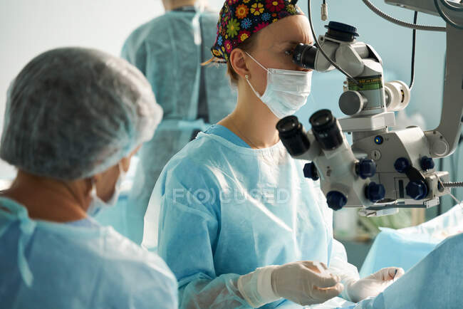 Medico donna adulto in maschera sterile e cappuccio medico ornamentale guardando attraverso il microscopio chirurgico contro il collega di coltura in ospedale — Foto stock