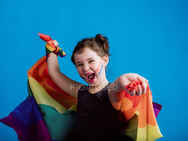 Lächelndes Mädchen mit aufgemalter Wange, das die Arme mit einer bunten Fahne auf leuchtend blauem Hintergrund in die Höhe reißt — Stockfoto