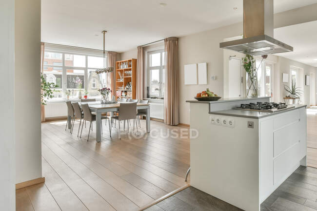 Convenient white kitchen island with stove and hood located in front of cozy dining area in modern open space apartment with white walls and ceiling and wooden floor — Stock Photo