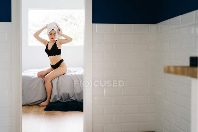 Schlanke Frau in weißem Handtuch-Turban und schwarzer Dessous sitzt nach dem Duschen zu Hause auf weichem Bett — Stockfoto