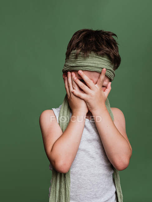 Anonymous shy karate boy in hachimaki headscarf covering face with hands on green background in studio — Stock Photo