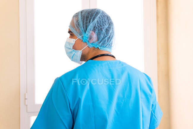 Vue arrière du médecin masculin masqué et uniforme debout près de la fenêtre dans la clinique — Photo de stock