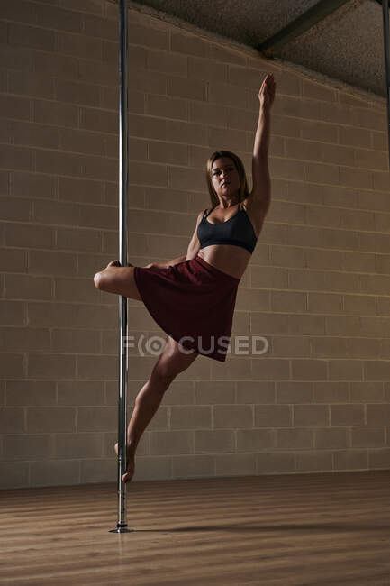 Slim female dancer in skirt and bra hanging on pole and performing in studio during rehearsal — Stock Photo