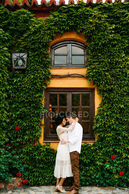 Vue latérale de couple multiethnique enchanté étreignant tout en se tenant sur le fond du bâtiment avec lierre vert croissant sur le mur — Photo de stock