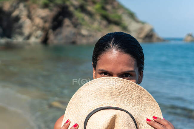 Hembra étnica turista oculta cara detrás de sombrero de paja mientras mira a la cámara contra el océano y la montaña a la luz del sol - foto de stock