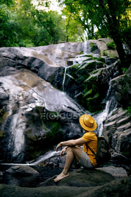 Vista lateral de un caminante masculino irreconocible sentado en la roca y admirando la cascada en el bosque - foto de stock
