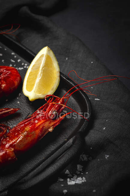 Delicious cooked red prawns on tray with coarse salt and juicy lemon pieces on dark background — Stock Photo