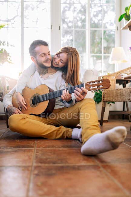 Cheerful tattooed male musician playing guitar near content female beloved while looking at each other in armchair in house room — Stock Photo