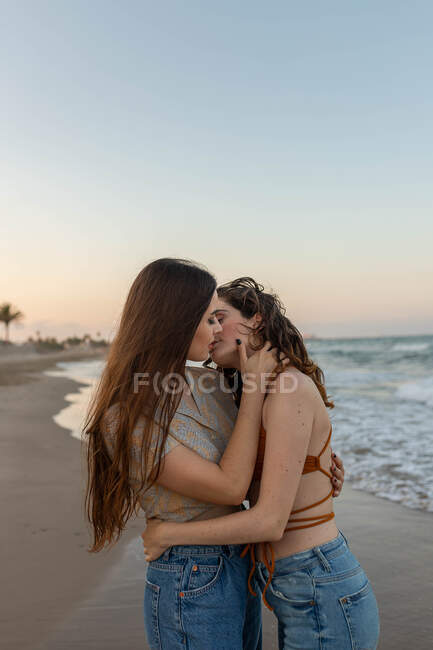 Young girlfriends embracing and kissing each other while standing on sandy beach near waving sea at sundown — Stock Photo
