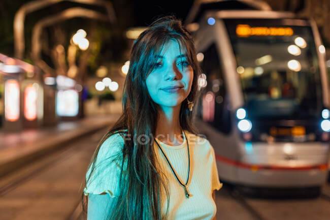 Lächelnde junge Frau mit langen Haaren blickt in der Abenddämmerung auf einer Stadtstraße mit Straßenbahnen in die Kamera — Stockfoto