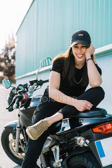 Motociclista feminina positiva sentada em moto estacionada na rua da cidade no dia ensolarado e olhando para a câmera — Fotografia de Stock