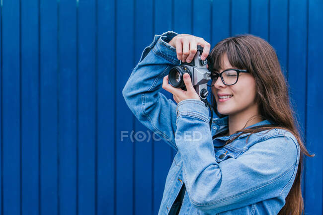 Side view of young female photographer in denim jacket taking picture on vintage photo camera on background of blue wall in city street — Stock Photo