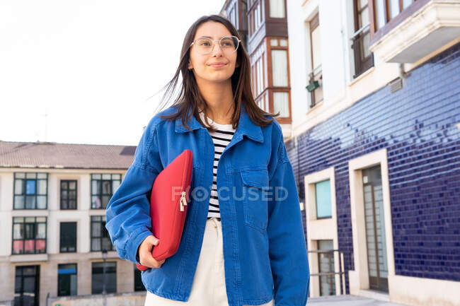 Alegre freelancer femenina con netbook en estuche rojo de pie en la calle de la ciudad y mirando hacia adelante - foto de stock