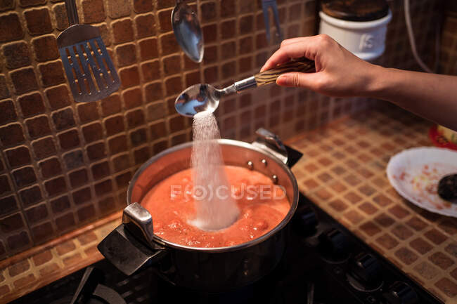 De arriba del cultivo femenino añadiendo la sal en la cacerola, mientras que cuecen la salsa marinara de los tomates a la estufa en la cocina - foto de stock