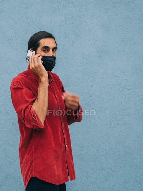 Calm ethnic male in protective mask talking on mobile phone while standing against blue wall in city during coronavirus — Stock Photo