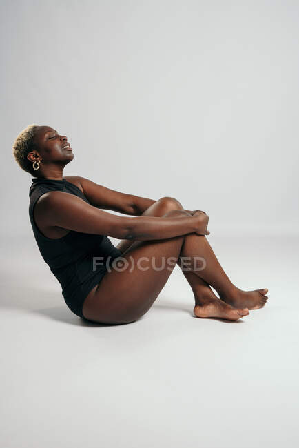 Афроамериканка в черном телесном костюме и с изогнутым телом сидит со скрещенными ногами в студии на сером фоне и смотрит в сторону — стоковое фото