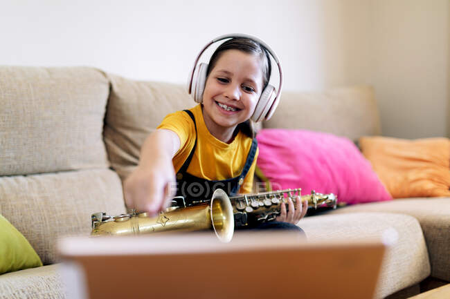 Niño consciente en auriculares y saxofón en el sofá grabación de vídeo en el teléfono celular en casa - foto de stock