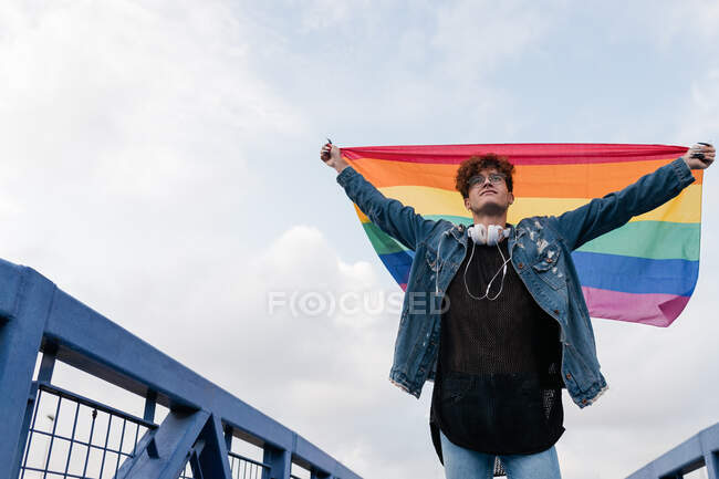 Desde abajo de los hombres homosexuales serios de pie con la bandera del arco iris en brazos levantados en el puente y mirando hacia otro lado - foto de stock