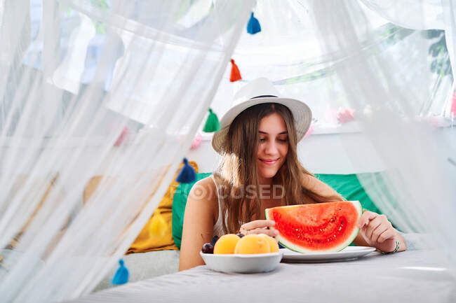 Positiva joven hembra sentada a la mesa con jugosa sandía madura y disfrutando del verano en la tienda del patio trasero - foto de stock