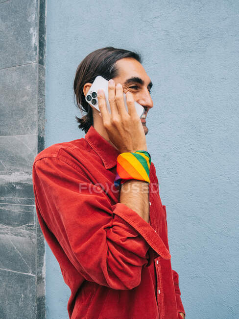 Delighted ethnic gay male with rainbow bandana on hand speaking on cellphone while standing near gray wall in city and looking away — Stock Photo