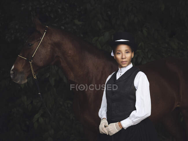 Señora adulta afroamericana confiada en ropa elegante y sombrero de pie con caballo marrón mientras mira la cámara cerca de los árboles durante el día - foto de stock